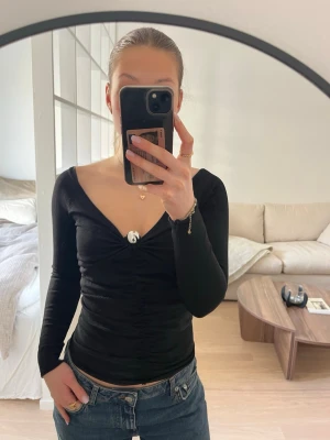 Svart v-ringad långärmad topp - Snygg svart långärmad topp med v-ringning och rynkade detaljer framtill. Sitter tight och har en smickrande passform. En silvrig detalj vid bröstet vilket ger en mer festlig känsla. Endast använd 1 gång och är i mycket fint skick. 