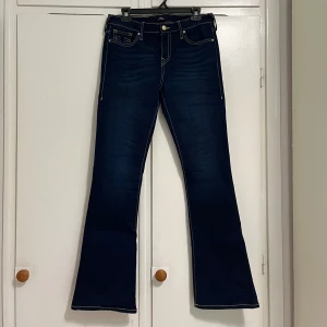 True Religion Bootcut Jeans Midwaist - Snygga mörkblå jeans från True Religion, modell Becca med bootcut och midwaist. Klassiska kontrastsömmar och ikoniska bakfickor med broderad logga. Tillverkade i slitstarkt jeansmaterial, perfekt för dig som gillar retrovibbar och statement-plagg.