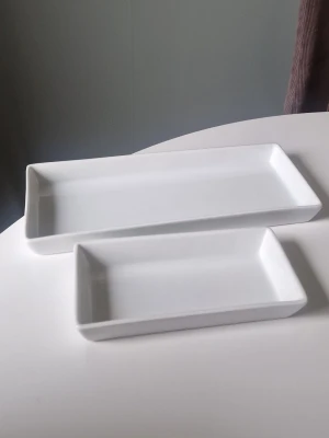 Fat - Säljer två stilrena vita rektangulära fat i porslin. Perfekta för servering av snacks, tapas eller som uppläggningsfat. Enkel och modern design som passar in i de flesta kök och hem. Båda faten har höga kanter och slät yta. De ska båda tåla värme, men kan spricka vid för snabba temperatur-skillnader. Det stora fatet tål mer värme än den lilla. Repor och märken finns på undersidan då de är använda. Inga övriga defekter eller märken.