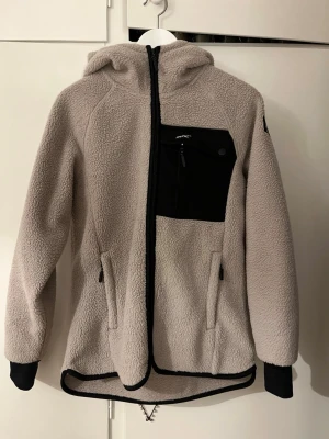 Beige teddyjacka med huva från Revolution race  - Mysig beige teddyjacka med svart bröstficka, dragkedja framtill och två sidofickor med dragkedja. Jackan har huva, svarta muddar vid ärmsluten och svarta detaljer längs kanterna. Perfekt för kalla dagar och riktigt skön att bära.