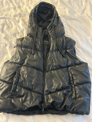 Svart väst med huva från Zara - Snygg svart väst från Zara med stor vadderad huva och dragkedja framtill. Västen har två fickor . Materialet är glansigt och ger en modern look. Perfekt för lager på lager och streetstyle. Är storlek L men sitter som S/M