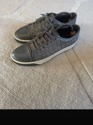 Lanvin - Snygga grå sneakers med krokodilpräglat mönster på sidorna och slät tå i mörkgrått. Skorna har vita sulor och matchande grå skosnören. Perfekta för dig som gillar en stilren men ändå unik look.