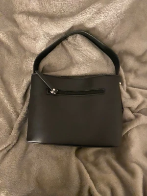 Svart stilren handväska i skinnimitation - Snygg svart handväska i skinnimitation med enkel, stilren design. Väskan har en bred axelrem och en dragkedjeficka på framsidan för småsaker. Perfekt för dig som gillar minimalistisk look och vill ha en väska som funkar till det mesta. OBBS! En silvrig knopp på vardera sida har lossnat, därav priset💕