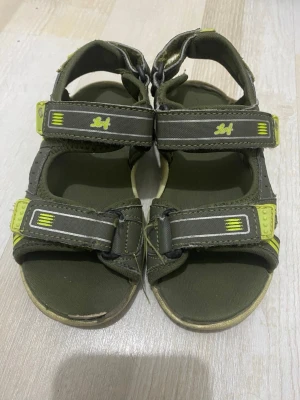 Gröna sportiga sandaler med kardborre och ljus - När man går blir det ljus. Säljer ett par gröna sportiga sandaler, justerbara kardborreband. Sandalerna har öppen tå och robust sula, perfekta för sommarens äventyr. Materialet är en mix av textil och syntet, vilket gör dem både lätta och bekväma.