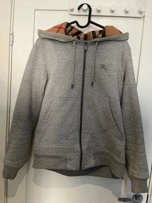 Grå hoodie från Burberry med dragkedja - Säljer en grå hoodie från Burberry med dragkedja framtill och klassiskt rutigt mönster i huvan. Haft i cirka 1 år säljer då jag tröttnat på den. Självklart äkta och Kvitto finns!