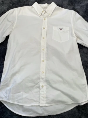 Vit skjorta från Gant med knappkrage - Klassisk vit skjorta från Gant med button-down krage och bröstficka med broderad logga. Skjortan har långa ärmar, knappar hela vägen och är gjord i mjuk bomull. Perfekt till jeans eller chinos för en clean look.