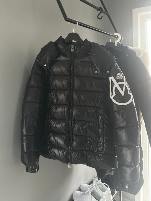 Svart dunjacka från Moncler - Snygg svart dunjacka från Moncler med blank finish och stor vit Moncler-logga på ena ärmen. Jackan har huva, dragkedja framtill och två sidofickor med dragkedja. Perfekt för kalla vinterdagar och riktigt trendig look.