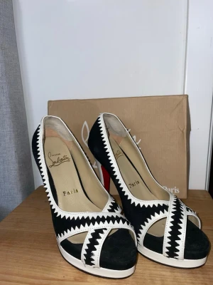 Svarta och vita pumps från Louboutin - Snygga pumps från Christian Louboutin i svart mocka med vita sicksack-detaljer och öppen tå. Skorna har hög vit klack, platåsula och den klassiska röda sulan. Perfekt för dig som gillar statement skor och exklusiv design.