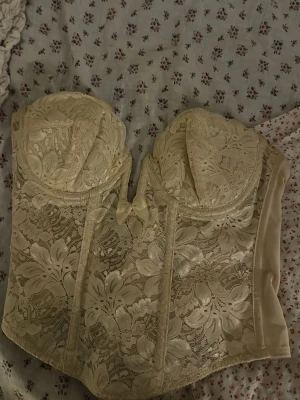 Korsette vintage  - Säljer en krämvit korsett med vackert blommigt spetsmönster och vadderade kupor. Korsetten har en söt rosett framtill och markerade sömmar för extra form. Perfekt för dig som vill ha en romantisk och elegant look.