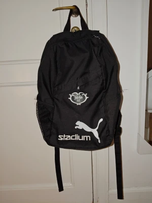 Svart Puma ryggsäck Järla IF - Snygg svart ryggsäck från Puma med vit Järla IF-logga och Stadium-tryck på framsidan. Ryggsäcken har ett stort huvudfack, ytterfack med dragkedja och sidoficka i mesh. Praktisk bärögla upptill och justerbara axelband. Perfekt för träning eller skola.  Ryggsäcken är tvättat och är redo för nästa ägare. Kommer från Rökfritt och djurfritt hem