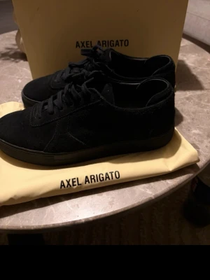 Svarta sneakers från Axel Arigato - Stilrena svarta sneakers från Axel Arigato i mocka med matchande svarta skosnören och sula. Klassisk låg modell som passar till det mesta och har diskret logga på sidan.