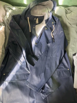 Mörkblå regnjacka med huva - Snygg mörkblå regnjacka med vit dragsko i huvan och tryckknappar framtill. Jackan har stora fickor och är tillverkad i ett vattenavvisande material med matt finish. Perfekt för regniga dagar och enkel att matcha med olika outfits. Har L och M i storlek