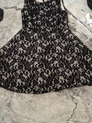 Svart spetsklänning från Kimi Mada - Säljer en svart kortklänning från Kimi Mada med vackert spetsmönster och figurnära överdel. Klänningen har ett elegant mönster i svart och vitt, är ärmlös och har en lätt utställd kjol. Tillverkad i polyester och elastan för skön stretch.