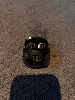 JBL svarta trådlösa in-ear hörlurar - Snygga svarta trådlösa in-ear hörlurar från JBL med transparent laddningsetui. Hörlurarna har en modern design med stilrena detaljer och silikonproppar för bra passform. Perfekta för musik, poddar och samtal på språng.