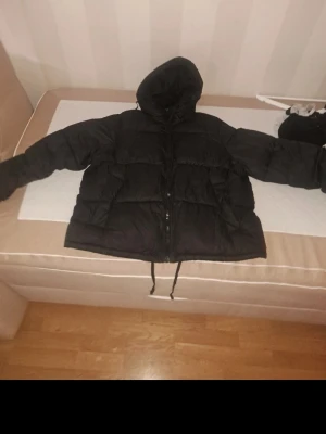 Svart pufferjacka med huva - Säljer en svart pufferjacka med huva och dragkedja framtill. Jackan har breda, vadderade paneler och dragsko nedtill för extra värme. Perfekt för kalla dagar och riktigt skön att bära. Klassisk modell som passar till det mesta.