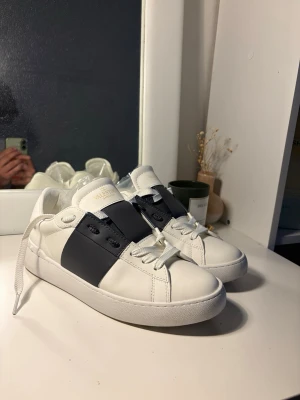 Vita Valentino sneakers med svart detalj - (Priset går att diskuteras) Snygga vita sneakers från Valentino med bred svart läderdetalj över snörningen och coola nitar på hälen. Klassisk låg modell med vita skosnören och diskret logga på plösen. Perfekta för dig som gillar stilrena och exklusiva sneakers.