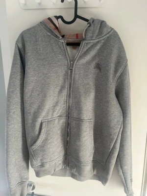 Grå zip hoodie från Burberry - Klassisk grå hoodie från Burberry med dragkedja framtill och huva. Ikoniskt rutigt foder i huvan och diskret Burberry-logga på bröstet. Två stora fickor framtill och ribbade muddar vid ärmslut och nederkant. Perfekt för en chill och stilren look.