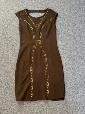 Svart klänning med guldiga och silvriga nitar samt öppen rygg - Snygg svart kortklänning med silver och guldfärgade nitar i geometriskt mönster framtill. Klänningen har öppen rygg och har en tight passform. Den är ärmlös och är perfekt för dig som vill sticka ut lite extra på en fest tex. Oanvänd utan defekter, fläckarna är bara damm då den inte kommit till användning. 