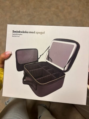 Sminkväska med spegel, uppladdningsbar - Praktisk svart sminkväska med inbyggd spegel och flera fack för smink och tillbehör. Väskan är uppladdningsbar och har måtten 26x23x11 cm. Perfekt för att organisera dina skönhetsprodukter och enkel att ta med tack vare handtaget.