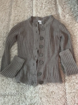 Grå stickad kofta med stora knappar - Mysig grå stickad kofta med stora matchande knappar framtill. Koftan har ett vågigt ribbmönster, lång ärm och rundad halsringning. Perfekt för lager på lager och enkel att styla till olika outfits.