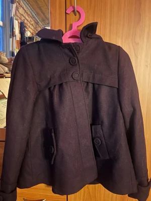 Svart/lila kappa med huva och stora knappar - Stilren svart/lila kappa från soaked med huva och stora dekorativa knappar framtill. Kappan har fickor med lock och knappar samt en lösare passform som ger en avslappnad look. Perfekt för kyligare dagar och enkel att matcha med olika outfits. Pris kan diskuteras vid snabb affär.