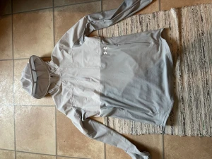 Ljusgrå hoodie/jacka från Under Armour - Snygg ljusgrå hoodie från Under Armour med dragkedja vid halsen och stor huva. Tillverkad i ett mjukt funktionsmaterial som andas. Har långa ärmar med diskret logga på mudden och Under Armour-logga på bröstet. Perfekt för träning eller chill.