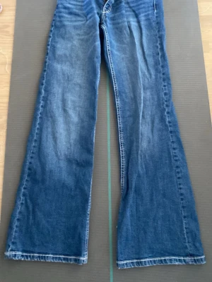 Blå bootcut jeans med broderade fickor - Säljer ett par blå bootcut jeans med snygg slitning och broderade bakfickor med vita detaljer. Jeansen har låg midja och vida ben, perfekta för en avslappnad och trendig look. Tillverkade i klassisk denim med knapp- och dragkedjegylf.