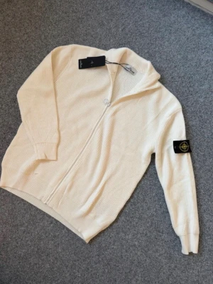 Vit ziptröja från Stone Island M - Snygg vit ziptröja från Stone Island med ribbad struktur och ikonisk svart patch med gul logga på ärmen. Tröjan har hög krage, långa ärmar och dragkedja framtill. Perfekt för dig som gillar streetwear och vill ha något stilrent.