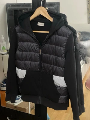 Svart hoodie med reflexdetaljer - Snygg svart hoodie från Moncler med quiltad front och reflexdetaljer på fickorna. Dragkedja framtill och huva för extra stilpoäng. Ärmar och rygg i mjukt tyg, medan framsidan har en glansig, vadderad look. Perfekt för dig som vill sticka ut.