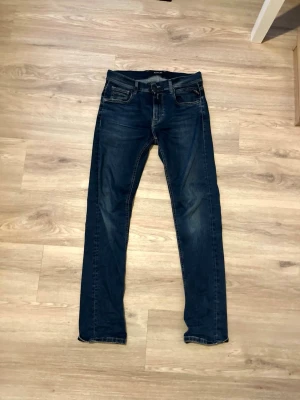 Replay anbass jeans - Snygga mörkblå slim fit jeans från Replay med klassisk femficksdesign och diskreta slitningar. Jeansen har smal passform och är tillverkade i ett mjukt denimtyg som sitter bekvämt. Perfekta för dig som gillar en stilren och modern look. Obs litet hål vid rumpan som knappt syns (bild om så önskas)
