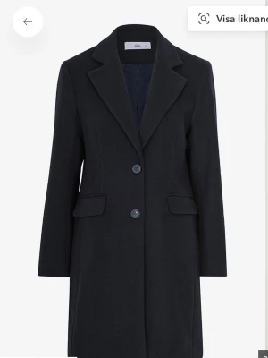 Svart klassisk kappa från Ellos - Stilren svart kappa från Ellos med två knappar framtill och klassisk krage. Kappan har två fickor med lock och är helfodrad för extra komfort. Perfekt för dig som gillar en tidlös och elegant look. Materialet ser ut att vara ullblandning.