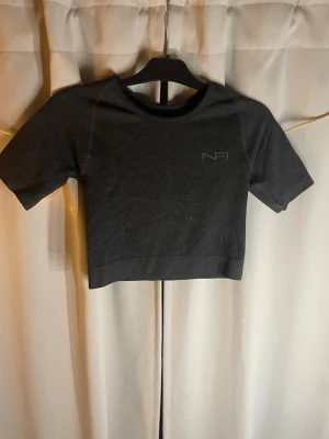 Mörkgrå croppad topp från NFA - Snygg mörkgrå croppad topp från NFA med korta ärmar och diskret logga framtill. Tillverkad i ribbad stretchig syntet som sitter tight och passar perfekt till träning eller chill. Enkel och stilren design som är lätt att matcha.