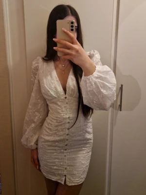 Vit kort klänning med puffärmar - Säljer en vit kort klänning med v-ringning, knappar framtill och stora puffärmar. Klänningen har ett subtilt blommigt mönster och är figurnära med en liten slits längst ner. Perfekt för dig som vill ha en trendig och elegant look.