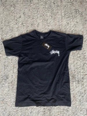 Stüssy x Tom & Jerry svart t-shirt - Svart t-shirt från Stüssy med Tom & Jerry-tryck på ryggen och vit Stüssy-logga på bröstet. T-shirten har korta ärmar och är gjord i mjuk bomull. Perfekt för dig som gillar streetwear och coola prints.