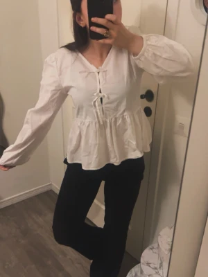 Vit blus med knyt och volang - Söt vit blus med långa puffärmar, knytning framtill och volang nedtill. Blusen har en avslappnad passform och är gjord i ett lätt tyg som ger en luftig känsla. Perfekt att styla med jeans eller kjol för en trendig look.