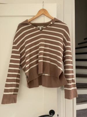 Randig brun och vit stickad hoodie - Säljer en stickad hoodie från WALK i brunt och vitt med breda ränder. Tröjan har en avslappnad passform, V-ringning och huva. Den har asymmetrisk nederkant och långa ärmar. Perfekt för en chill och trendig look.