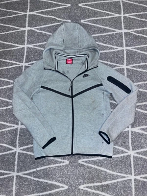 Ljusgrå Nike hoodie med dragkedja - Säljer Nike Tech Hoodie i toppskick.         Har använt ett par gånger, inte ofta.    Den har inga fläckar eller märken!     Köpt för 1399kr på XXL!                             Levererar snabbt✅