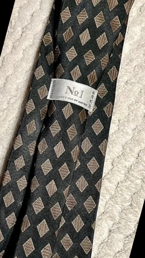 Svart slips med rutmönster från No1 - Snygg svart slips från No1 med guld rutmönster. Tillverkad i ett glansigt material som ger en stilren look. Perfekt accessoar för att lyfta din outfit med en unik touch.