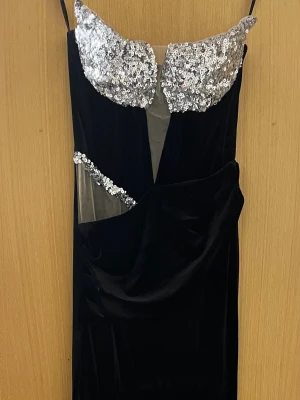 Svart långklänning med paljettdetaljer - Elegant svart långklänning med tunna axelband och glittriga silverpaljetter över bysten och sidan. Klänningen har en unik cut-out detalj och är tillverkad i mjuk sammet som ger en lyxig känsla. Perfekt för dig som vill sticka ut på festen.