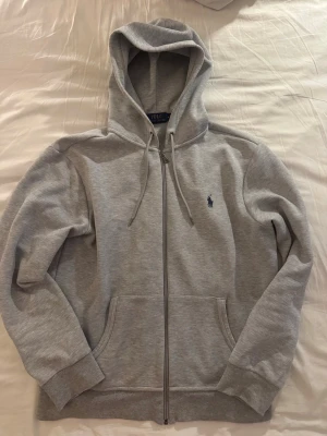 Grå zip hoodie från Polo Ralph Lauren - Snygg grå hoodie med dragkedja från Polo Ralph Lauren. Klassisk design med huva och dragsnören, två fickor fram och den ikoniska lilla loggan broderad på bröstet. Tillverkad i mjukt sweatshirtmaterial som är riktigt skönt.