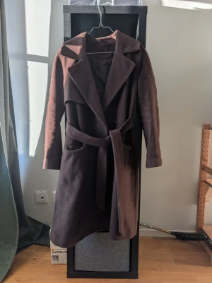 Brun lång kappa med skärp - Stilren brun kappa i ullmix med klassisk krage och knytskärp i midjan. Kappan har två fickor framtill och en mjuk, borstad yta som ger en lyxig känsla. Perfekt för kyliga dagar när du vill vara både varm och snygg.