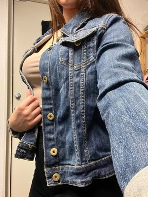 Klassisk blå jeansjacka - Snygg blå jeansjacka med guldfärgade knappar och klassiska sömmar. Jackan har två bröstfickor med lock och knapp. 