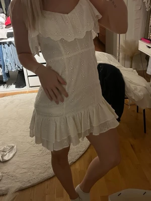 Vit kort klänning med volang - Söt vit kort klänning med broderade detaljer och volangkant både upptill och nedtill. Klänningen har smala axelband och är ärmlös, perfekt för sommarens alla tillfällen. Banden är uppsydda på klänningen jag är 168 och den passar mig i längden men man kan enkelt sprätta bort sömnen om man vill ta bort de uppsydda. Klänningen har en liten smink fläck inuti men inget som syns