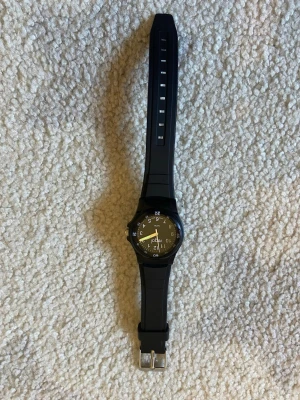 Svart klocka från Regal med gummiband - Stilren svart klocka från Regal med rund urtavla och tydliga vita siffror. Klockan har ett svart gummiband och silverfärgat spänne. Perfekt för dig som vill ha en enkel och klassisk accessoar till vardagslooken.