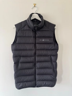 Svart väst från Sail Racing - Snygg svart dunväst från Sail Racing i bra skick. Västen har två sidofickor med dragkedja och logga på bröstet. Perfekt för användning nu på våren!