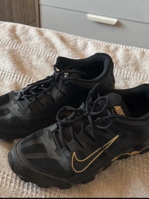 Nike Reax 8 Tr strl 45,5 - Ett av par svarta träningsskor från Nike med guldfärgade detaljer. Skorna har snörning framtill och en robust sula.  Har varsitt hål på ensidig av vardera skor, kanske går att laga för den som orkar!  Enda skavankerna jag hittar är de små hålen på skorna, samt ett annat litet hål, annars vad jag kan se bara smutsiga och lite slitna på under sulan och inuti. 