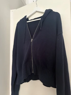 Mörkblå stickad hoodie med dragkedja - Säljer en mörkblå stickad hoodie med dragkedja framtill och ribbade muddar. Hoodien har en avslappnad passform, huva med dragsko och något kortare. Perfekt för en chill och stilren look.