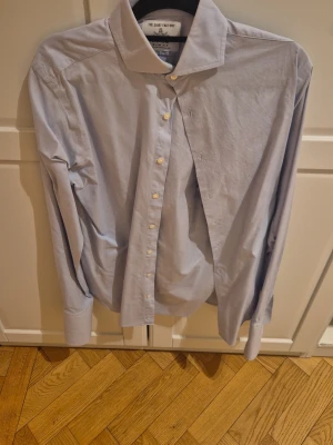 Ljusblå skjorta från The Shirt Factory - Klassisk ljusblå skjorta från The Shirt Factory i slim fit-modell. Skjortan har långa ärmar, vit knappstängning och en stilren krage. Perfekt för dig som gillar en clean och enkel look. Dubbelvikt manschetter för manschettknappar 