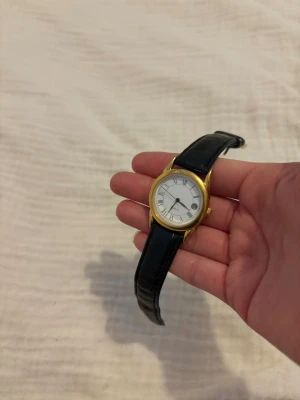 Klassisk guldfärgad klocka med äkta läder band - Stilren klocka med guldfärgat runt boett och svart äkta läderband med krokopräglat mönster. Urtavlan är vit med romerska siffror och datumvisning. Perfekt accessoar för dig som gillar tidlös design. Så himla fin och kom tyvärr inte till användning