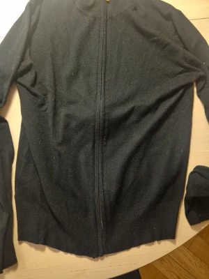 Svart ziptröja från William de Faye - Klassisk svart ziptröja från William de Faye. Tröjan har hel dragkedja framtill, ribbade muddar och är tillverkad i mjukt stickat material. Perfekt för lager på lager och enkel att matcha med olika outfits.